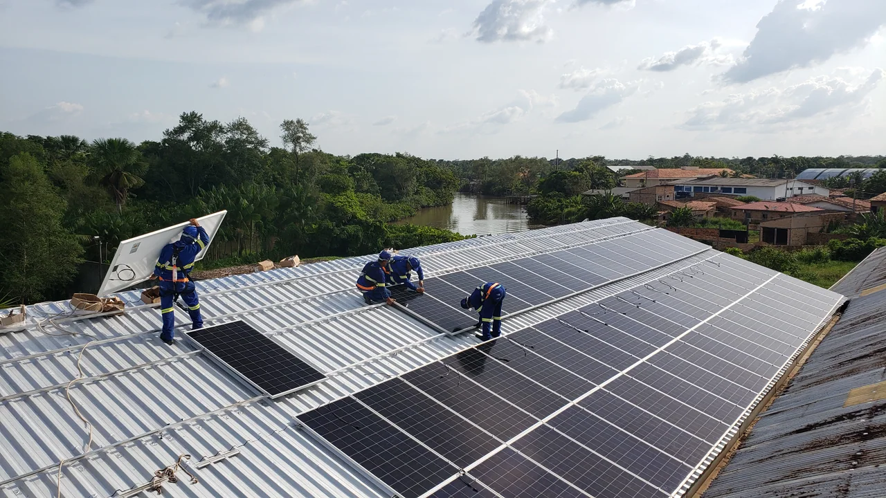 Equipe instala painéis solares no telhado de um edifício; ao fundo aparecem casas e vegetação de Abaetetuba, no Pará.