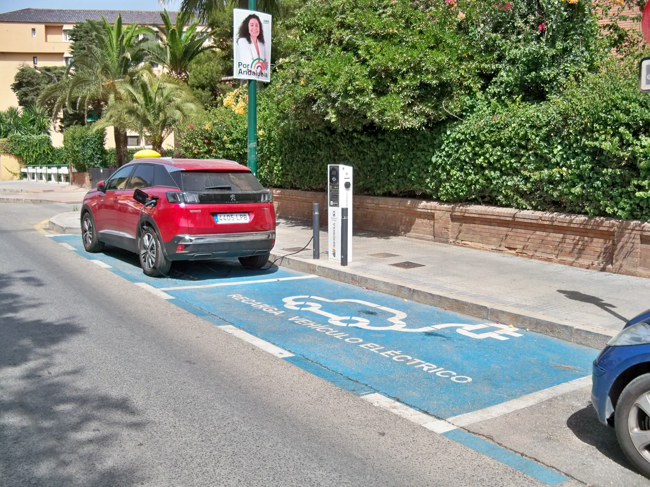 Vehículo rojo conectado por cable a un punto de recarga Iberdrola en una plaza pública azul de Málaga; fotografía de archivo de 2022.