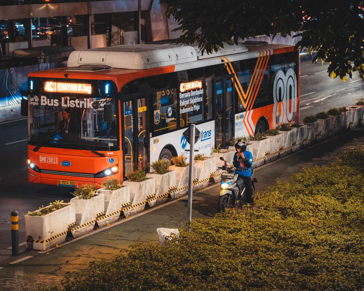 Ônibus elétrico laranja e branco da TransJakarta circula à noite em Jacarta, com uma motocicleta ao lado.